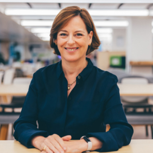 Smiling woman in a dark blue blouse sits at a desk in a bright office environment.