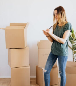 Young woman stands beside stacked cardboard moving boxes, taking notes on a clipboard.