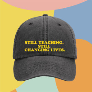 Close-up of a gray cap with bold yellow text reading 'STILL TEACHING. STILL CHANGING LIVES.' set against a colorful abstract background.