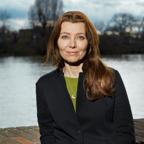 Portrait of a woman with long brown hair wearing a dark blazer and green top, standing by a lake with trees in the background.