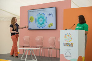 Two women present on a stage: one points to a large screen with a diagram titled 'A Whole-School Approach to RSHE' behind them, pink and orange backdrops visible, empty chairs in front, and a white podium to the right with a colorful 'Festival of Education' logo behind it.