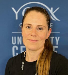 Portrait of a staff member with long hair in a black shirt, wearing a staff lanyard and badges, against a blue university backdrop.