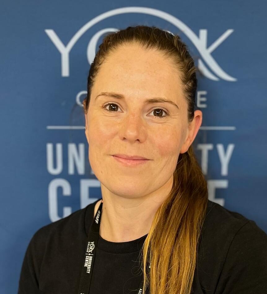 Portrait of a staff member with long hair in a black shirt, wearing a staff lanyard and badges, against a blue university backdrop.