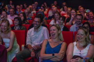Audience members in a theater laughing and enjoying a performance on red seats in the foreground and others clapping behind them.