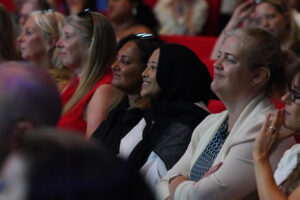 Diverse group of women seated in a conference hall, smiling and watching a presentation.