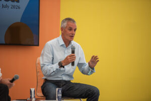 Man in a light blue shirt speaks into a handheld microphone during a panel against a bright yellow backdrop.