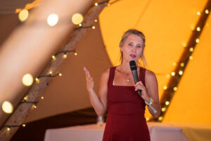 Woman in a red dress speaks into a handheld microphone at a festive indoor event with warm yellow lights in the background.