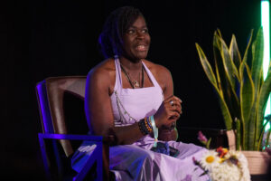 A woman wearing a lavender dress sits on a stage chair, speaking or answering questions, with decorative plants and flowers nearby under stage lighting.