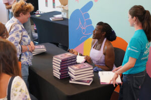 Author signing copies at a table during a book signing event; stacked books, attendees waiting, and a colorful backdrop.