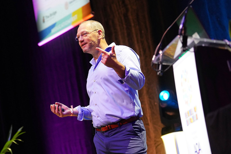 Speaker on stage wearing a light blue shirt with a headset mic, gesturing with his hands while presenting at a conference.