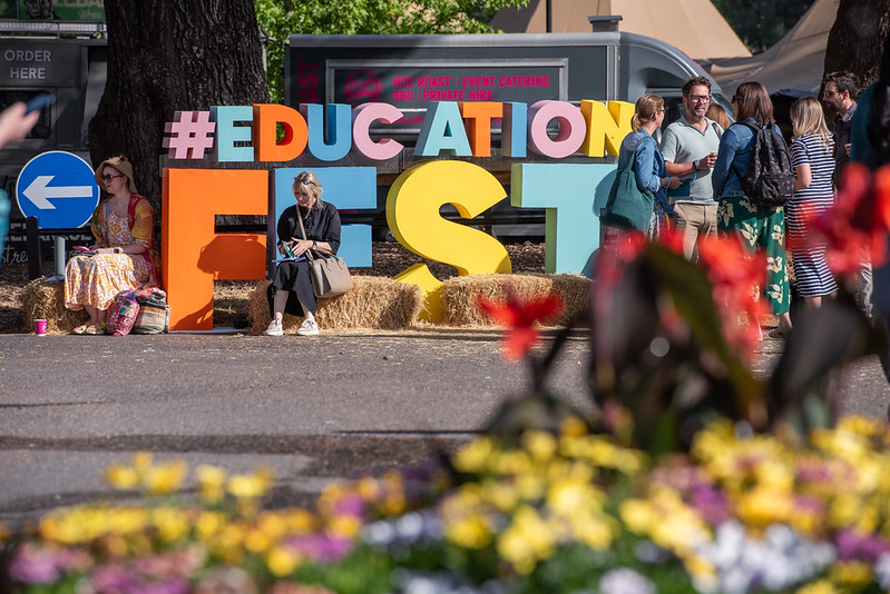 Colorful letters spelling '#EDUCATION FEST' at an outdoor festival, with people chatting nearby and flowers in the foreground.