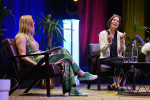 Two women sit on a stage in conversation; left wears a green patterned dress, right wears a cream cardigan, with plants and colorful curtains behind them.