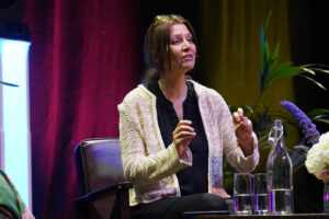 Woman in a cream knit cardigan and black top seated on stage, gesturing as she speaks, with water bottles and plants nearby.