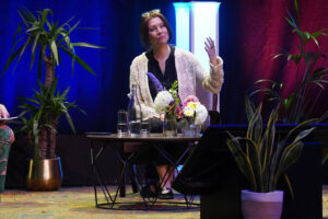Woman seated at a low table on a stage, speaking and gesturing with one hand, surrounded by potted plants and a floral arrangement.