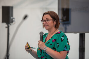 Woman in a green patterned dress speaks into a handheld microphone during a presentation, holding a remote in one hand.