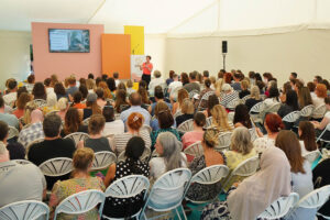 Speaker presenting to a large audience in a white tent, with a screen and colorful stage panels behind.