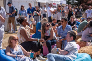 Diverse group lounging on hay bales at an outdoor festival, chatting and drinking under tents at the background?