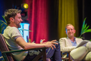 Two men sit on stage in conversation: the left speaker with a headset laughs and gestures, the right man in a light suit smiles.