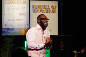 Man with beard and glasses on stage, speaking at the Festival of Education with banners in the background.