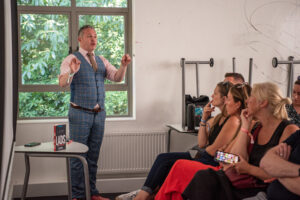 Man in a blue plaid suit gives a talk to a small audience in a bright room by a window with green foliage outside.