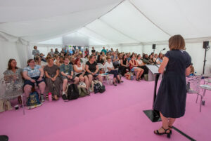 Woman speaker at a podium addressing a seated audience inside a white tent with a pink floor.",