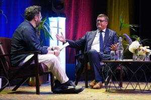 Two men in a stage interview sit in armchairs; one in a dark suit and glasses gestures while the other holds papers.