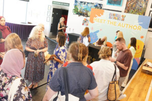 People gather around a table labeled 'Meet the Author' at a festival, with an artistically painted backdrop and attendees waiting or purchasing books.