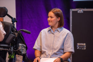 Woman in a wheelchair on a stage, wearing a light blue striped shirt and holding papers, looking to the side against a purple backdrop.