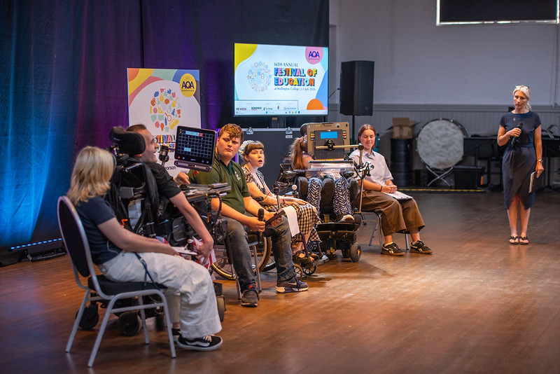 People in wheelchairs on a stage at a Festival of Education, with a presenter standing to the right and event branding on screens in the background.