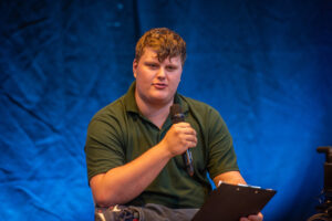 Man in a green polo speaks into a handheld microphone on a stage, holding a clipboard with a blue backdrop behind him.