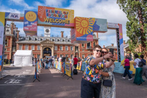 Couple takes a selfie at the Festival of Education entrance under a colorful 'Welcome' arch with a brick campus building behind them.