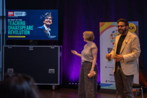 Two speakers stand on a stage beside a banner while a screen promotes the Teaching Shakespeare Revolution in the background.