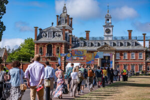 People form a long queue to enter a grand red-brick campus building with a clock tower on a sunny day, banners above the entrance.