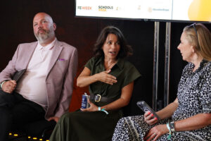 Three panelists seated on a stage during a discussion, with the central woman speaking and gesturing toward her chest; logos appear on a screen above.