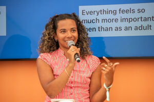 Woman speaks into a handheld microphone on stage, gesturing with her left hand.