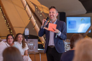 Man in a blue blazer speaks into a microphone, holding an orange card during a lit event or presentation in a hall.