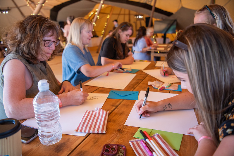 Group of people seated at a wooden table outdoors in a tent, drawing on white sheets with markers under string lights.