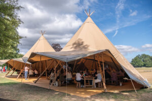 Giant beige teepee tents set up in a park with people seated at tables under a canopy, enjoying an outdoor gathering on a sunny day