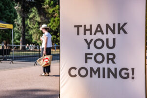 Large banner reads 'THANK YOU FOR COMING!' with a woman in a hat and a tote bag standing in the background at an outdoor event.