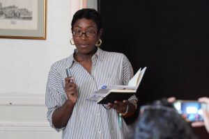 Woman with short hair and glasses, wearing a striped shirt, holding a notebook and pen while speaking to an audience.