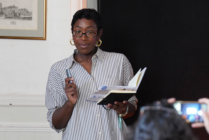 Woman with short hair and glasses, wearing a striped shirt, holding a notebook and pen while speaking to an audience.